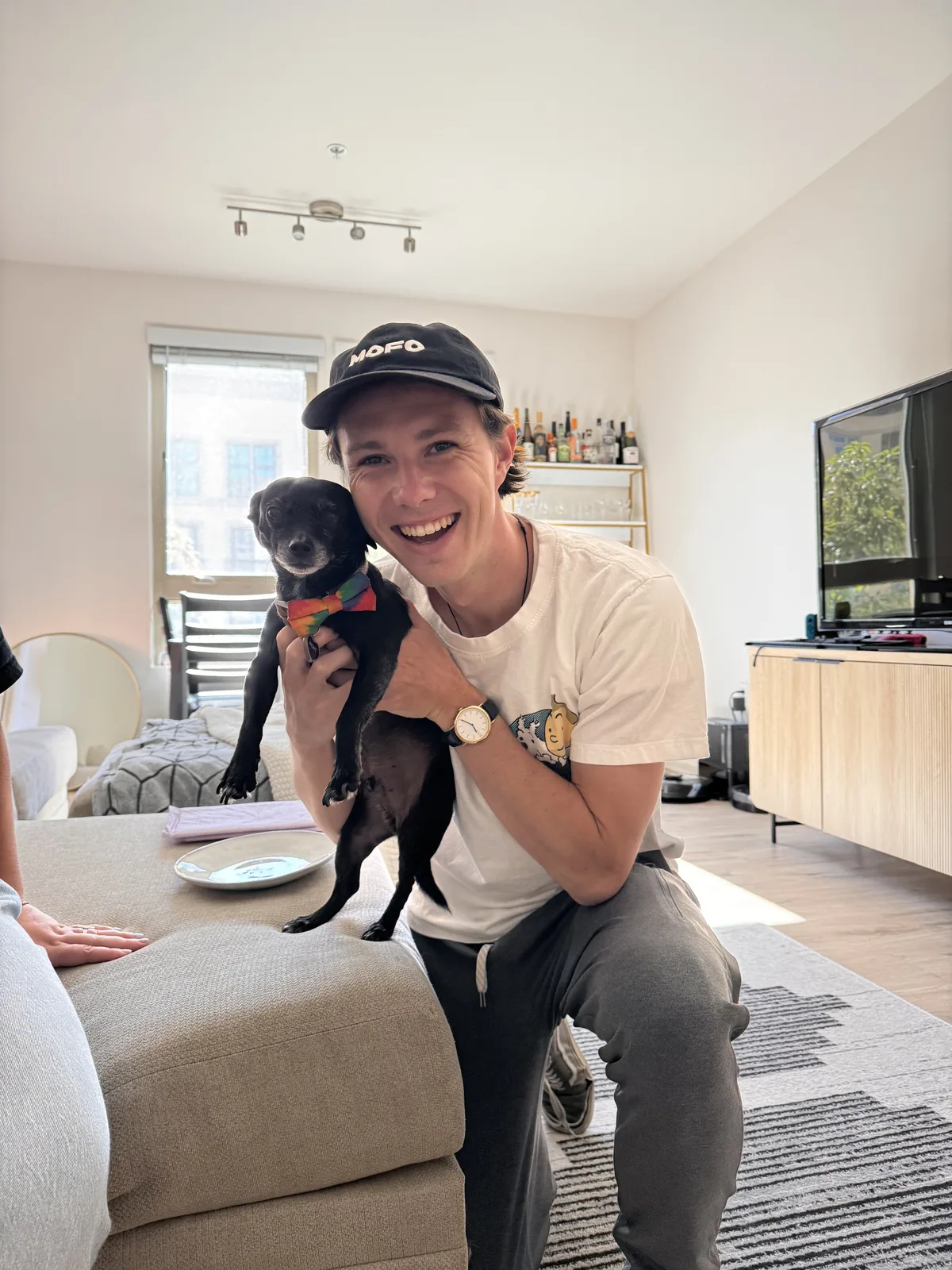Sam grinning while holding a small black dog wearing a rainbow bow tie