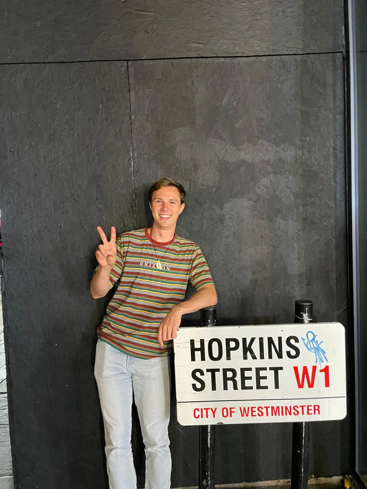 Sam giving a peace sign next to a Hopkins Street W1 street sign in London